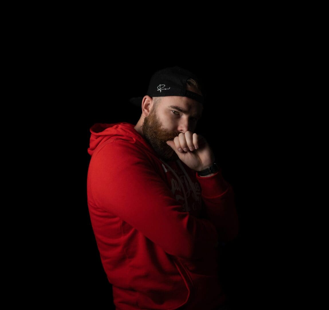 Man in red hoodie and cap, thoughtful pose against black background.